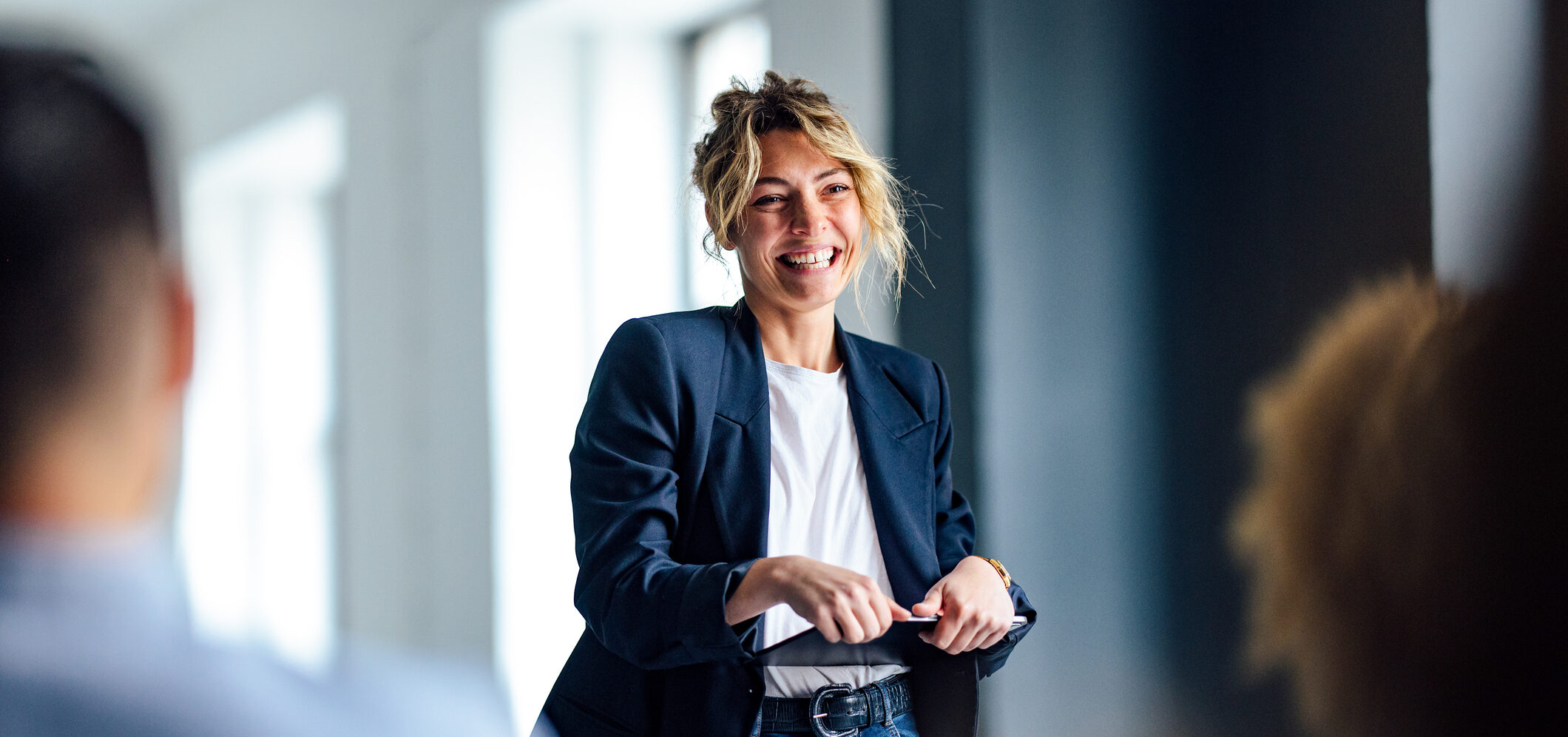 Beautiful cheerful woman in a dark blue jacket standing in front of a crowd looking down at a tablet.